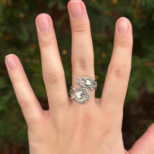 May include: A silver-toned bypass ring crafted from a repurposed spoon handle, featuring an open design with two ends that overlap. Each end displays an embossed heart motif surrounded by intricate scrollwork and a small floral detail.