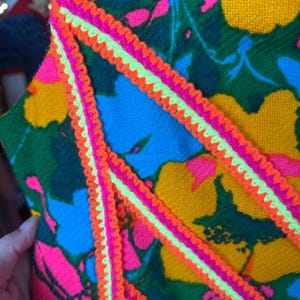 May include: A textile garment featuring a vibrant floral pattern in shades of yellow, green, blue, and hot pink. The fabric is accented with decorative zigzag trim in bright neon orange and lime green, applied in diagonal lines across the surface.