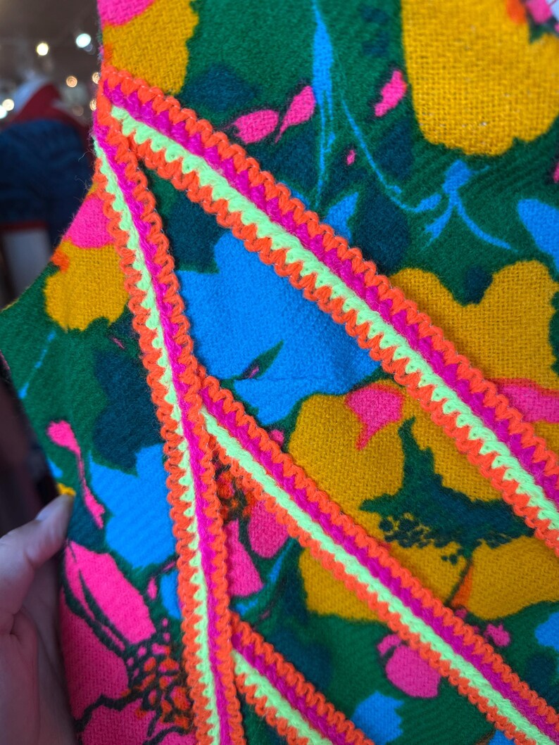 May include: A textile garment featuring a vibrant floral pattern in shades of yellow, green, blue, and hot pink. The fabric is accented with decorative zigzag trim in bright neon orange and lime green, applied in diagonal lines across the surface.