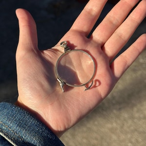 Puede incluir: Un monóculo plateado con montura circular y pequeños brazos curvos. El monóculo tiene un pequeño lazo en la parte superior para una cadena. El objeto se sostiene en una mano abierta.