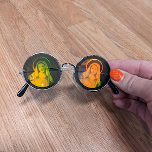 May include: Round sunglasses with silver frames and black temples. The lenses feature a colorful image of a woman with a halo. The sunglasses are held by a hand with orange nail polish.