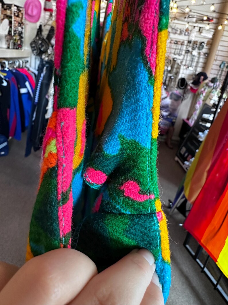 May include: A thick, felted fabric bag featuring a vibrant, abstract pattern in bright shades of hot pink, electric blue, forest green, and golden yellow. The material has a fuzzy, textured surface with visible stitched seams along the edges.