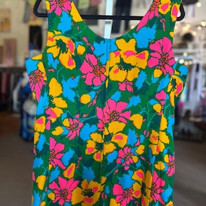 May include: A sleeveless dress featuring a vibrant floral pattern with bright pink, yellow, and light blue flowers set against a deep green background. The garment has a scoop neckline and a textured fabric finish, displayed on a black hanger.