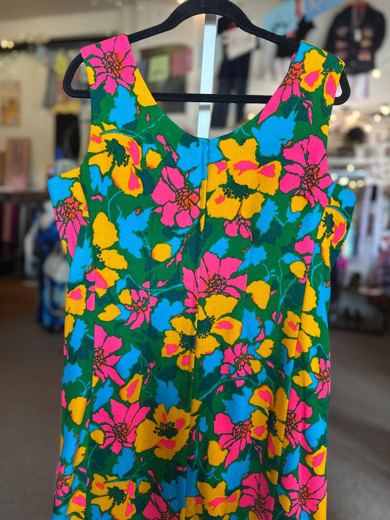 May include: A sleeveless dress featuring a vibrant floral pattern with bright pink, yellow, and light blue flowers set against a deep green background. The garment has a scoop neckline and a textured fabric finish, displayed on a black hanger.
