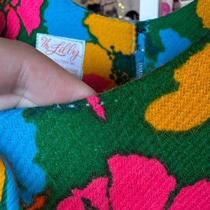 May include: A sleeveless dress featuring a vibrant floral pattern in hot pink, bright blue, and golden yellow on a green background. A white fabric label with the text The Lilly Pulitzer Inc is sewn inside the neckline near a blue zip.