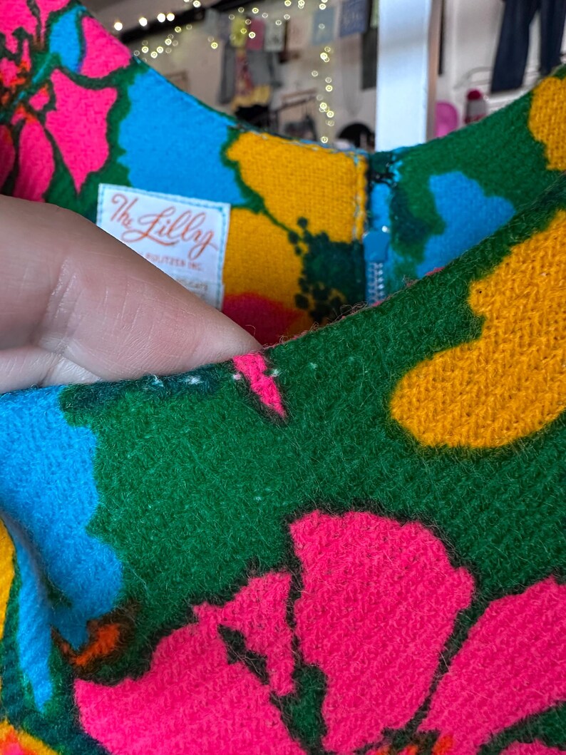 May include: A sleeveless dress featuring a vibrant floral pattern in hot pink, bright blue, and golden yellow on a green background. A white fabric label with the text The Lilly Pulitzer Inc is sewn inside the neckline near a blue zip.