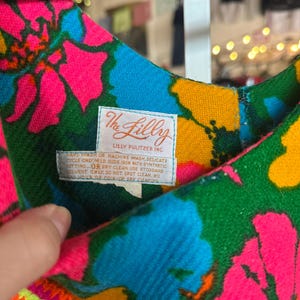 May include: A sleeveless dress featuring a vibrant floral pattern in shades of hot pink, bright blue, green, and yellow. A white fabric label is sewn into the neckline, displaying the text The Lilly Lilly Pulitzer Inc. and care instructions.