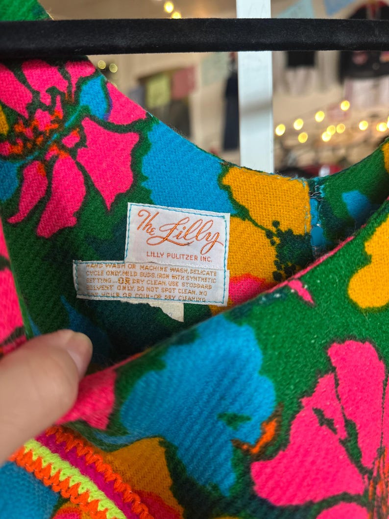 May include: A sleeveless dress featuring a vibrant floral pattern in shades of hot pink, bright blue, green, and yellow. A white fabric label is sewn into the neckline, displaying the text The Lilly Lilly Pulitzer Inc. and care instructions.