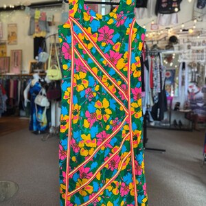 May include: A sleeveless midi dress featuring a vibrant floral pattern in shades of pink, yellow, and blue against a green background. The dress is accented with diagonal stripes of bright pink and yellow trim running across the front panel.