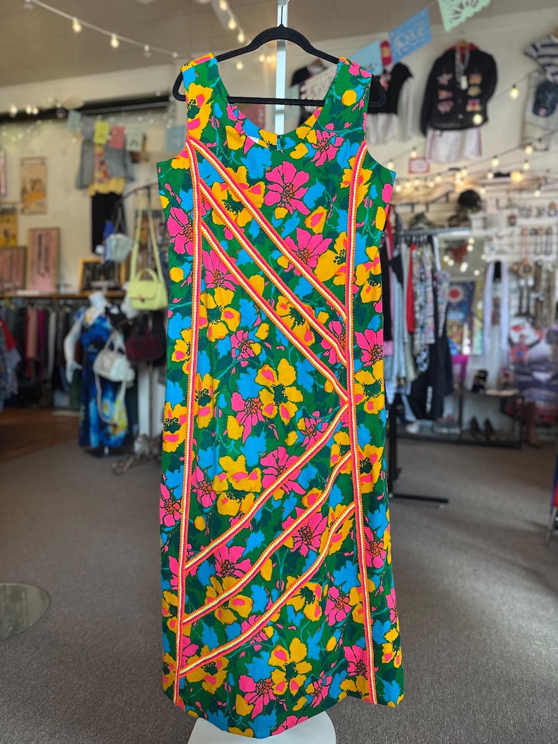 May include: A sleeveless midi dress featuring a vibrant floral pattern in shades of pink, yellow, and blue against a green background. The dress is accented with diagonal stripes of bright pink and yellow trim running across the front panel.