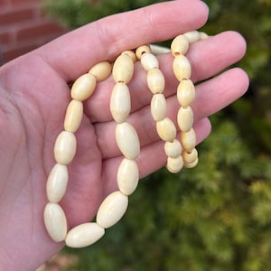 May include: A cream-coloured beaded necklace featuring oval-shaped beads. The necklace is held in a hand against a backdrop of green foliage and a brick wall. The beads are strung together, creating a simple, elegant design.