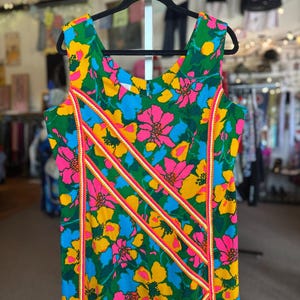 May include: A sleeveless dress featuring a vibrant floral pattern in shades of green, bright pink, yellow, and blue. The garment is accented with three diagonal stripes of orange and white trim that run across the front of the bodice.