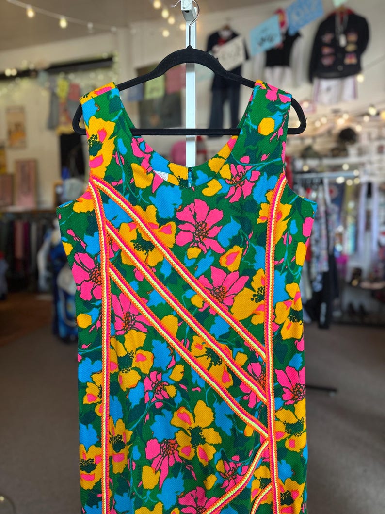 May include: A sleeveless dress featuring a vibrant floral pattern in shades of green, bright pink, yellow, and blue. The garment is accented with three diagonal stripes of orange and white trim that run across the front of the bodice.