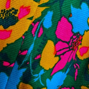 May include: A textile fabric featuring a vibrant floral pattern with bold, saturated colours. The design includes large, stylised flowers in shades of hot pink, mustard yellow, and bright sky blue, set against a deep forest green background.