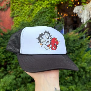 May include: A black and white trucker hat with a white front panel featuring a cartoon image of a woman with a red devil on her shoulder. The hat has a black mesh back panel.