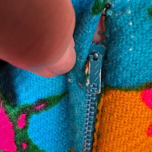 May include: A textured fabric garment featuring a vibrant, multi-coloured pattern of bright blue, orange, green, and hot pink. A metal zip with a teal-coloured pull tab is integrated into the seam, showing signs of wear on the metal finish.