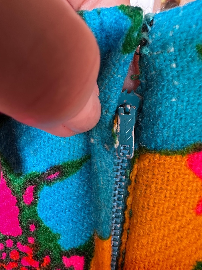 May include: A textured fabric garment featuring a vibrant, multi-coloured pattern of bright blue, orange, green, and hot pink. A metal zip with a teal-coloured pull tab is integrated into the seam, showing signs of wear on the metal finish.