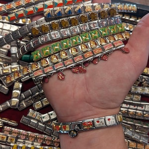 May include: A hand wearing a silver charm bracelet with various charms, including hearts, flags, and words like "Mom" and "Rodeo". The bracelet is surrounded by a variety of other charms in a display case.
