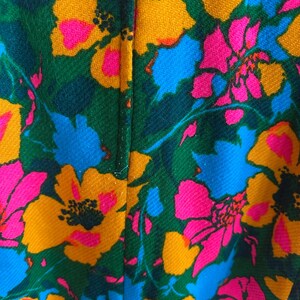 May include: A textile fabric featuring a vibrant floral pattern with large, stylised flowers in shades of bright yellow, hot pink, and sky blue, set against a deep forest green background. The material has a visible diagonal weave texture.