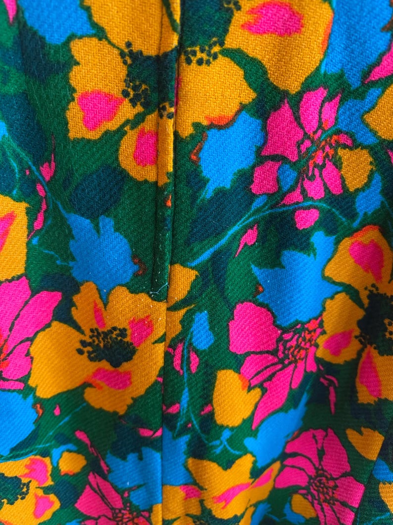 May include: A textile fabric featuring a vibrant floral pattern with large, stylised flowers in shades of bright yellow, hot pink, and sky blue, set against a deep forest green background. The material has a visible diagonal weave texture.