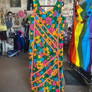 May include: A sleeveless knee-length dress featuring a vibrant floral pattern in shades of green, yellow, blue, and pink. The garment is accented with diagonal stripes of orange and white trim that run across the front of the fabric.