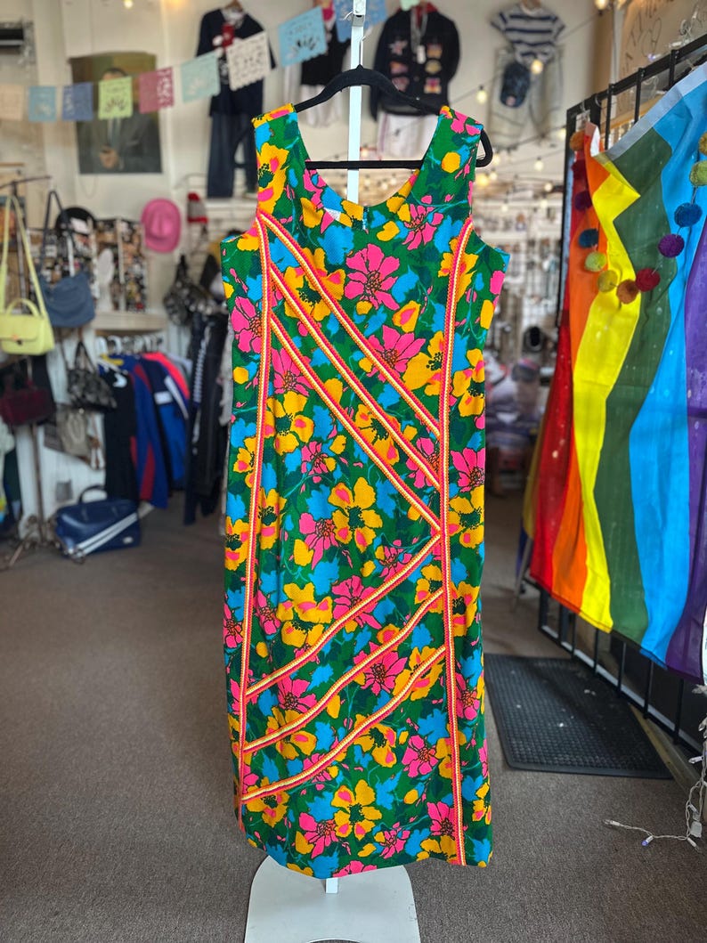 May include: A sleeveless knee-length dress featuring a vibrant floral pattern in shades of green, yellow, blue, and pink. The garment is accented with diagonal stripes of orange and white trim that run across the front of the fabric.