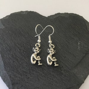 May include: Silver-coloured earrings with small seated goblin figures. The earrings have a hook design for pierced ears and are displayed on a dark grey, textured surface. The goblin figures are detailed with facial features and seated with their knees bent.