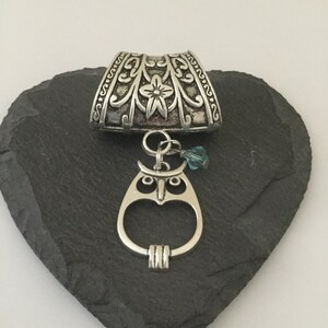 May include: Silver pendant with an ornate floral design and an owl charm hanging from it. The pendant is on a dark gray heart-shaped stone.