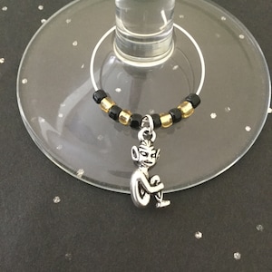 May include: A silver charm of a small, seated figure with large ears hangs from a black and gold beaded wine glass charm.