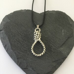 May include: A silver pendant necklace with a rope knot design. The necklace is on a black cord and is displayed on a heart-shaped slate.