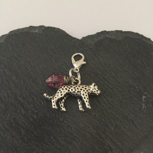 May include: A silver leopard charm with black spots, attached to a clasp. A pink crystal bead hangs from the clasp. The charm is set against a dark, textured background. The charm is suitable for a bracelet or necklace.