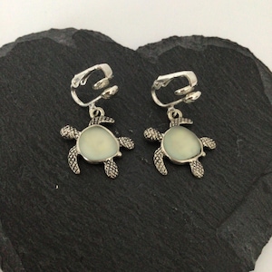 May include: A pair of silver-coloured clip-on earrings in the shape of sea turtles. Each earring has a pale green, oval centre and detailed turtle flippers. The earrings are shown on a dark, textured surface.
