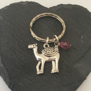 May include: A silver camel charm keyring with a small, dark pink crystal charm. The keyring is on a dark grey heart-shaped stone.