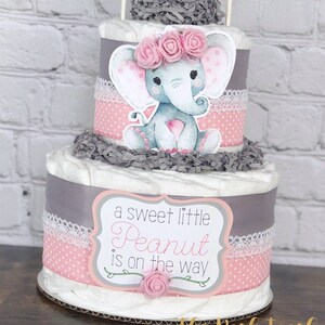 May include: A two-tier diaper cake decorated with a gray and pink elephant, pink flowers, and a sign that reads "a sweet little Peanut is on the way".
