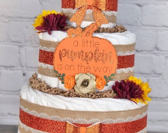 May include: A three-tier diaper cake decorated with burlap, orange and burgundy flowers, and orange glitter bows. The cake is topped with a pumpkin-shaped sign that reads "a little pumpkin is on the way".
