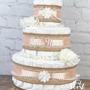 K&ouml;nnte beinhalten: Eine dreist&ouml;ckige Windeltorte, verziert mit Burlap-Band, Spitze und wei&szlig;en Blumen. Die Torte ist mit dem Namen "Rylee Marie" personalisiert.