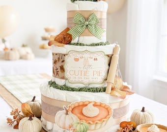 May include: A three-tiered diaper cake decorated with a fall theme, featuring a green gingham bow, a wooden rolling pin, and a sign that reads A little CUTIE PIE IS ON THE WAY. The base is adorned with a pumpkin pie illustration and small pumpkins.