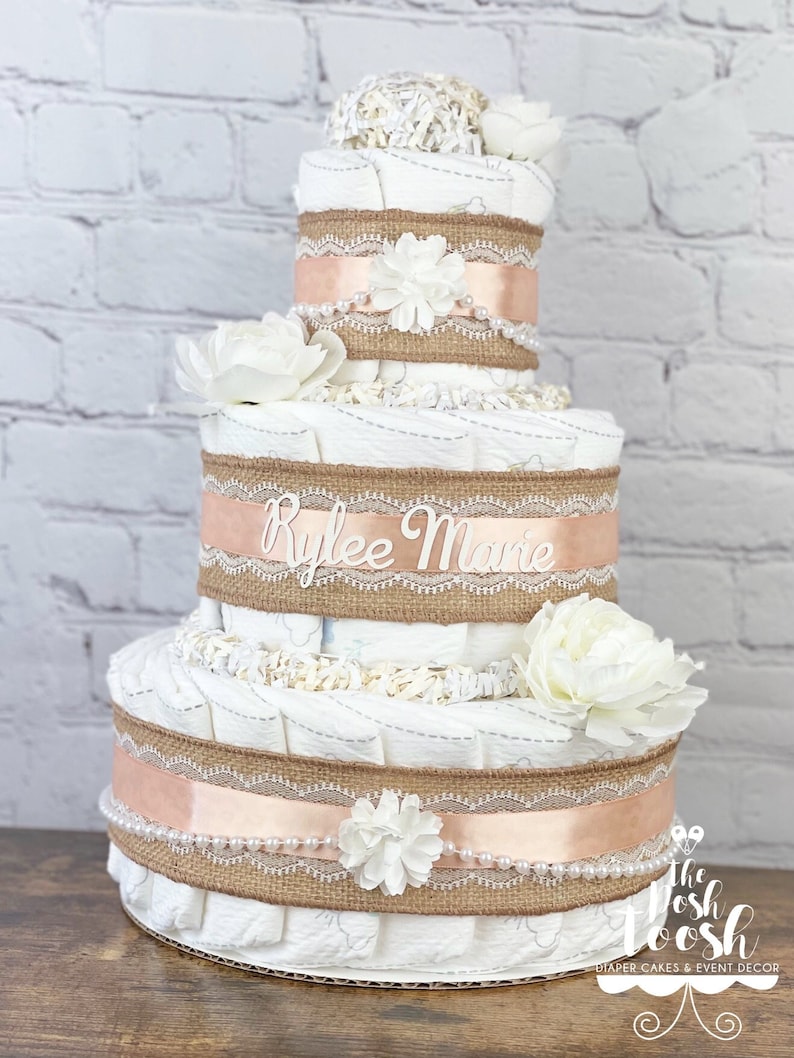 K&ouml;nnte beinhalten: Eine dreist&ouml;ckige Windeltorte, verziert mit Burlap-Band, Spitze und wei&szlig;en Blumen. Die Torte ist mit dem Namen "Rylee Marie" personalisiert.