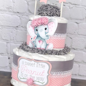 May include: Two-tiered diaper cake decorated with pink and gray ribbons, a gray elephant with pink polka dots, and a sign that reads "a sweet little Peanut is on the way".