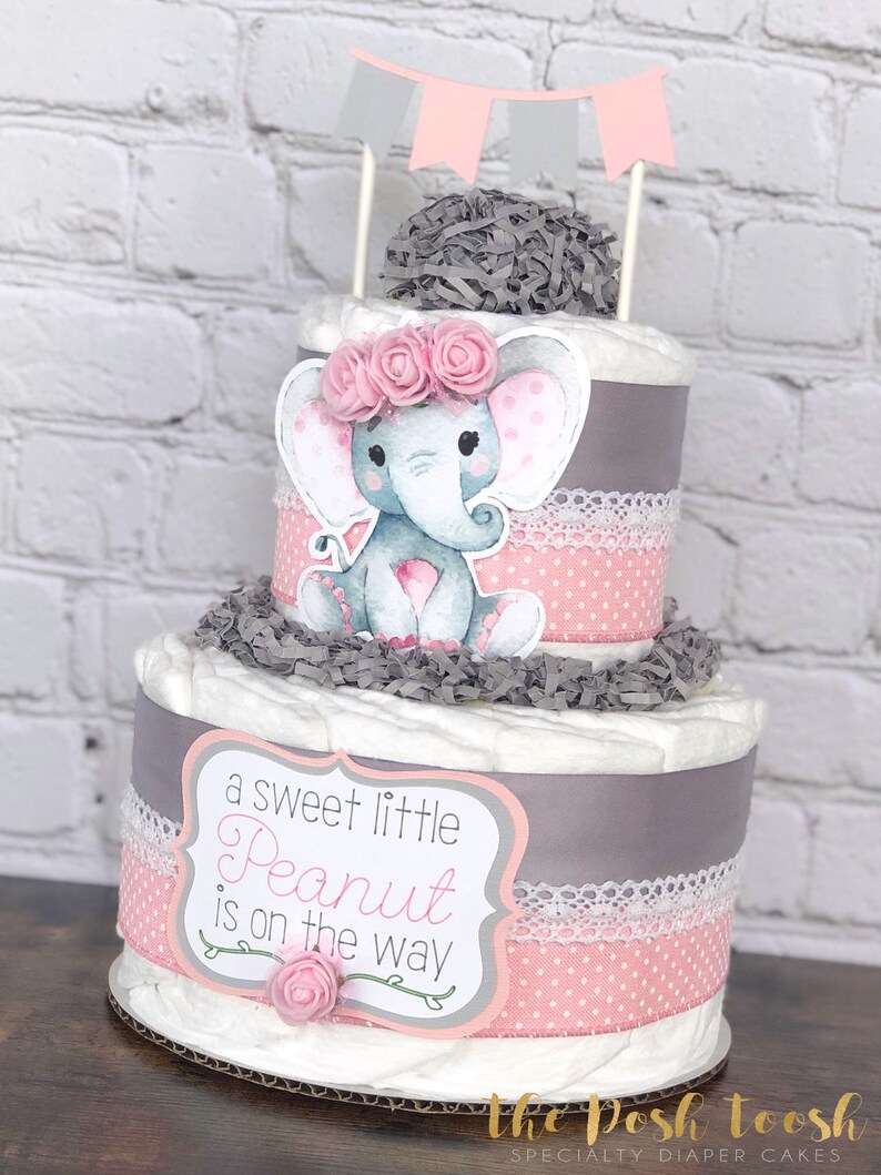 May include: Two-tiered diaper cake decorated with pink and gray ribbons, a gray elephant with pink polka dots, and a sign that reads "a sweet little Peanut is on the way".