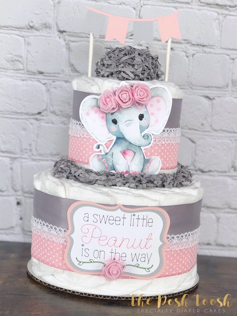 May include: A two-tier diaper cake decorated with a gray and pink elephant, pink flowers, and a sign that reads "a sweet little Peanut is on the way".
