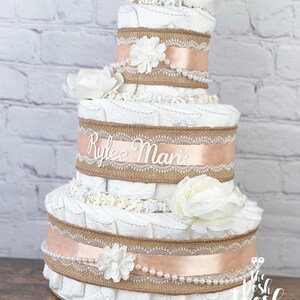 K&ouml;nnte beinhalten: Eine dreist&ouml;ckige Windeltorte, verziert mit Sackleinen, Spitze und einem pfirsichfarbenen Band. Die Torte ist mit wei&szlig;en Blumen und Perlen verziert. Die mittlere Etage hat ein wei&szlig;es Schild mit der Aufschrift "Rylee Marie".