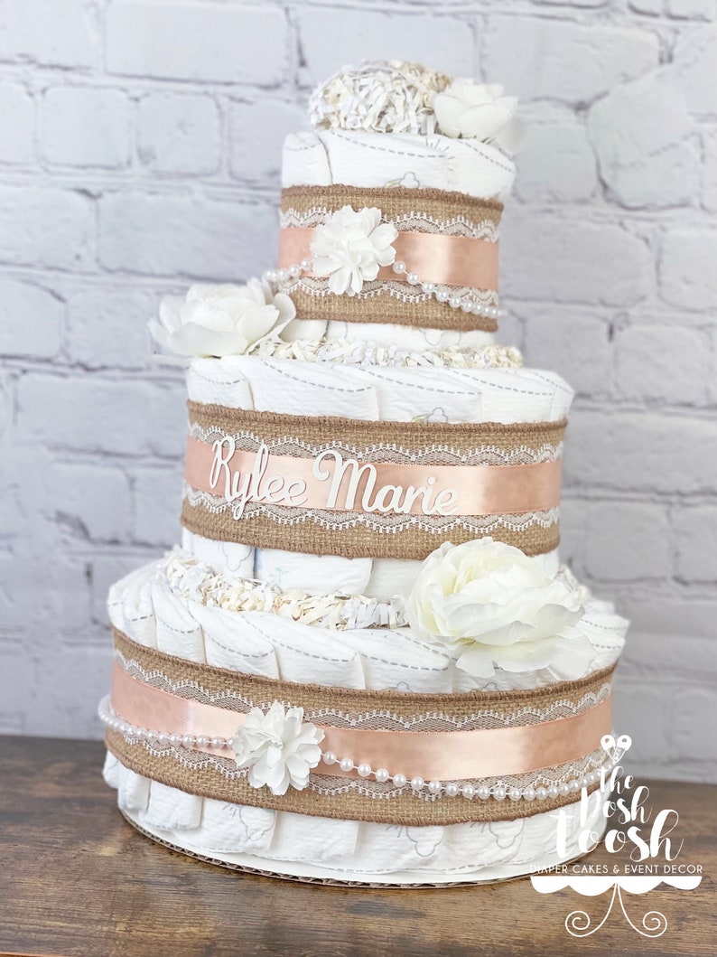 K&ouml;nnte beinhalten: Eine dreist&ouml;ckige Windeltorte, verziert mit Sackleinen, Spitze und einem pfirsichfarbenen Band. Die Torte ist mit wei&szlig;en Blumen und Perlen verziert. Die mittlere Etage hat ein wei&szlig;es Schild mit der Aufschrift "Rylee Marie".