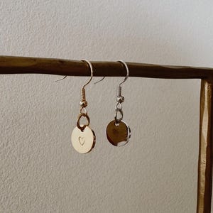 May include: A pair of gold and silver drop earrings. The gold earring features a small heart etched into the circular pendant. The earrings are displayed on a gold-coloured stand against a neutral background.