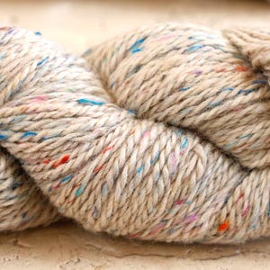 May include: Close-up of a skein of beige yarn with colorful flecks of blue, red, and pink. The yarn is thick and twisted, suggesting it's suitable for knitting or crocheting projects. The texture appears soft and slightly fuzzy.