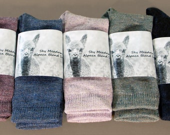 May include: Five pairs of alpaca blend socks in a variety of colours, including pink, blue, green, and black. Each pair is packaged with a label featuring a baby alpaca and the text "Sky Meadow Alpaca Blend Socks".