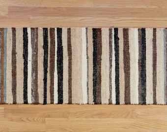 Puede incluir: Una alfombra tejida con un patrón a rayas en tonos marrones, beige y negros. La alfombra está hecha de fibras naturales y tiene un aspecto rústico y hecho a mano.