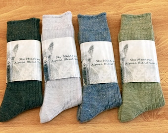 Puede incluir: Cuatro pares de calcetines de mezcla de alpaca en diferentes colores: verde, gris, azul y verde oliva. Cada calcetín tiene una etiqueta blanca con el texto "Sky Meadows Alpaca Blend Socks".