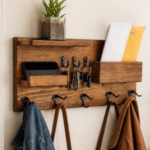 Könnte beinhalten: Ein hölzerner Eingangsorganisator mit einem Regal, Postfächern, Schlüsselhaken und Mantelhaken. Oben steht eine kleine Topfpflanze. Eine Jeansjacke und eine Ledertasche hängen an den Haken. Ein Smartphone und Schlüssel sind sichtbar.