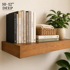 May include: A wooden floating shelf, 10-12 inches deep, displays books, a woven basket with a folded blanket, and a potted plant. The books have titles like "THE KINFOLK HOME" and "THIS IS HOME". The shelf is stained a warm brown.
